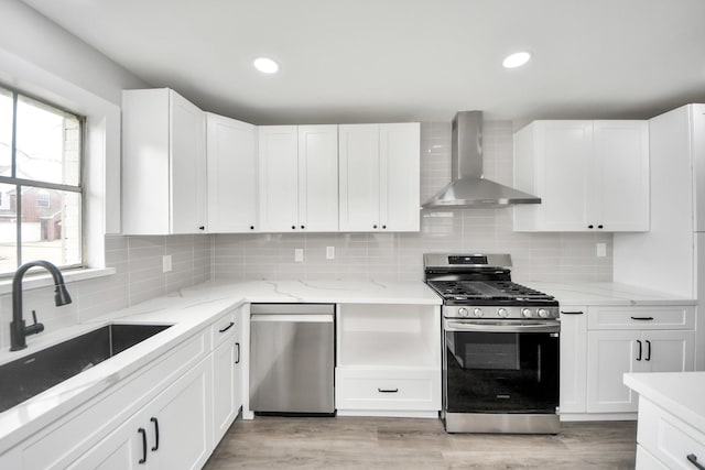 kitchen with light wood finished floors, wall chimney exhaust hood, a sink, stainless steel appliances, and backsplash