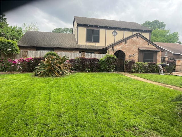 tudor house with brick siding, a shingled roof, fence, stucco siding, and a front yard