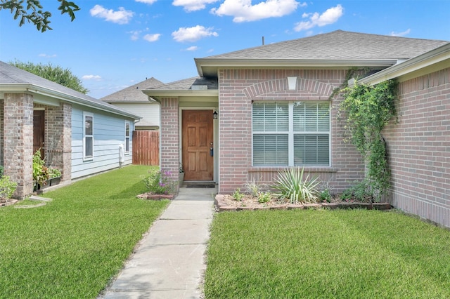 doorway to property featuring roof with shingles, fence, a lawn, and brick siding