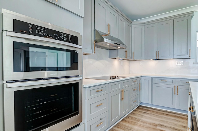 kitchen featuring double oven, black electric cooktop, under cabinet range hood, light countertops, and light wood finished floors