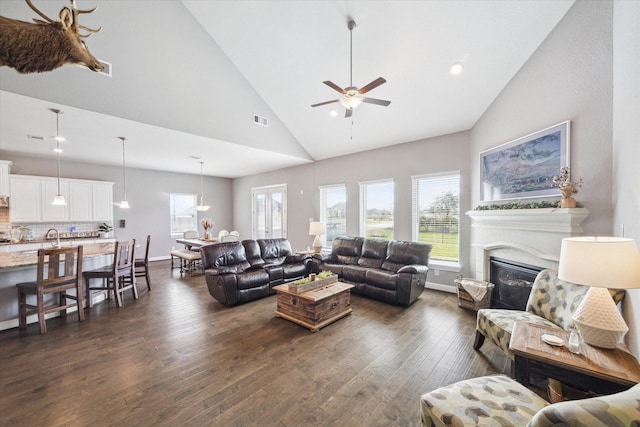 living area featuring dark wood-style floors, visible vents, a glass covered fireplace, high vaulted ceiling, and baseboards