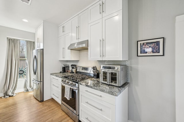 kitchen with under cabinet range hood, stainless steel appliances, light wood-type flooring, dark stone counters, and tasteful backsplash