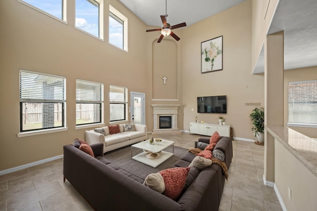 living area featuring a glass covered fireplace, a towering ceiling, light tile patterned floors, baseboards, and ceiling fan