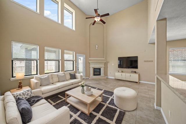 living area with a ceiling fan, a tiled fireplace, a high ceiling, light tile patterned floors, and baseboards