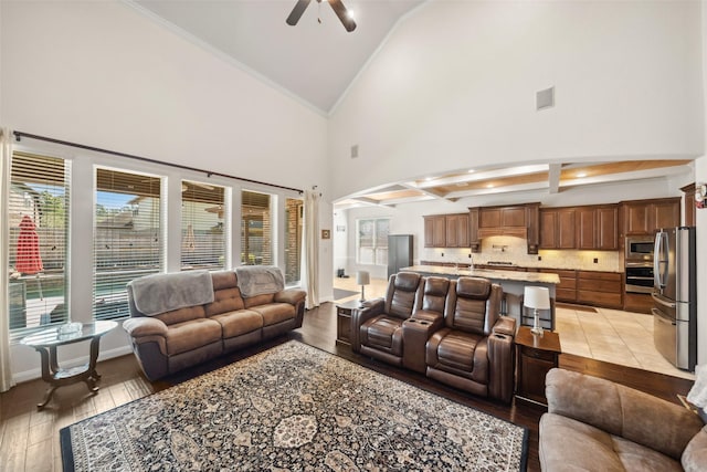 living room featuring light wood finished floors, a ceiling fan, high vaulted ceiling, beamed ceiling, and baseboards