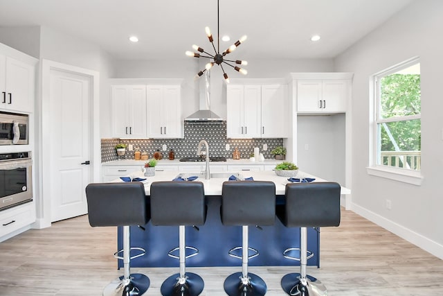 kitchen with tasteful backsplash, light countertops, appliances with stainless steel finishes, an island with sink, and wall chimney exhaust hood