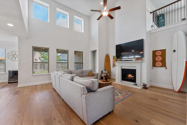 living room with a ceiling fan, beverage cooler, light wood-type flooring, a warm lit fireplace, and baseboards