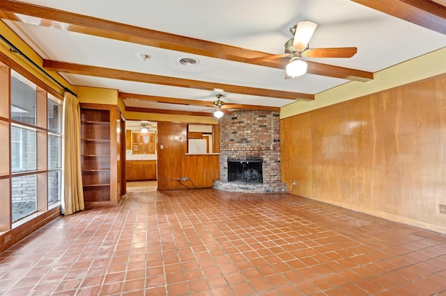 unfurnished living room featuring wooden walls, visible vents, a brick fireplace, ceiling fan, and beam ceiling