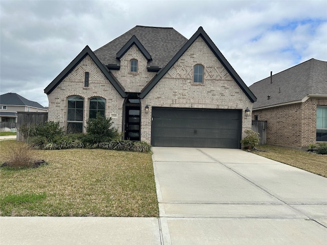 french country style house with a shingled roof, a front yard, concrete driveway, and brick siding