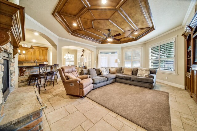 living room featuring arched walkways, coffered ceiling, ornamental molding, a stone fireplace, and ceiling fan with notable chandelier