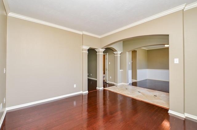 empty room featuring arched walkways, decorative columns, ornamental molding, wood finished floors, and baseboards