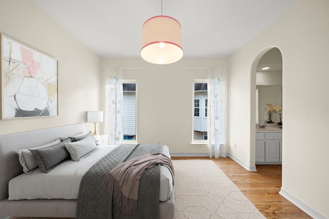 bedroom with light wood-type flooring, arched walkways, baseboards, and ensuite bathroom