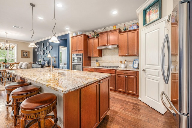 kitchen with under cabinet range hood, a sink, ornamental molding, appliances with stainless steel finishes, and brown cabinets