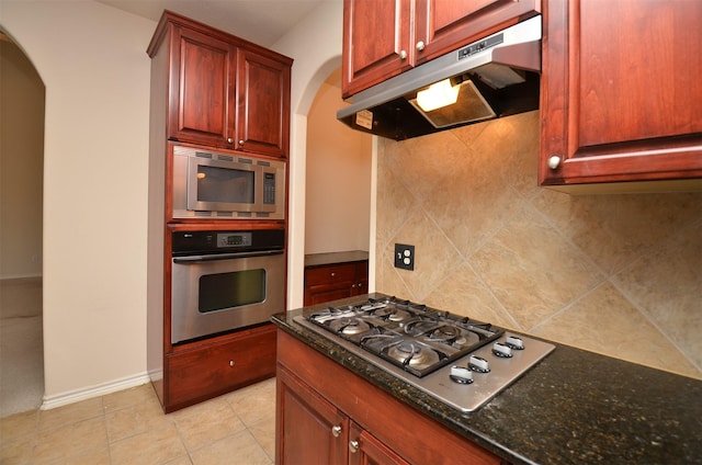 kitchen featuring dark stone counters, arched walkways, stainless steel appliances, under cabinet range hood, and backsplash