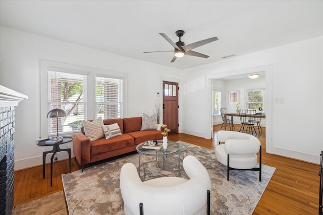 living area featuring baseboards, wood finished floors, a brick fireplace, and ceiling fan