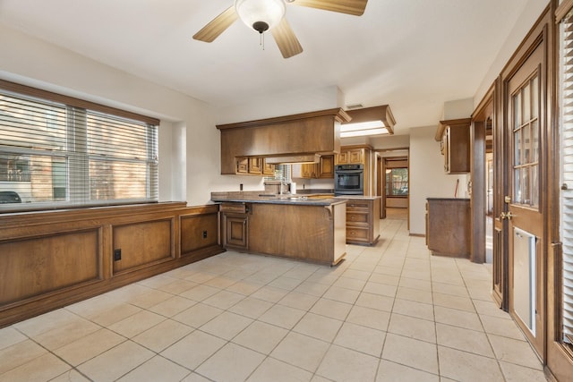 kitchen featuring light tile patterned floors, a peninsula, a ceiling fan, black oven, and brown cabinets