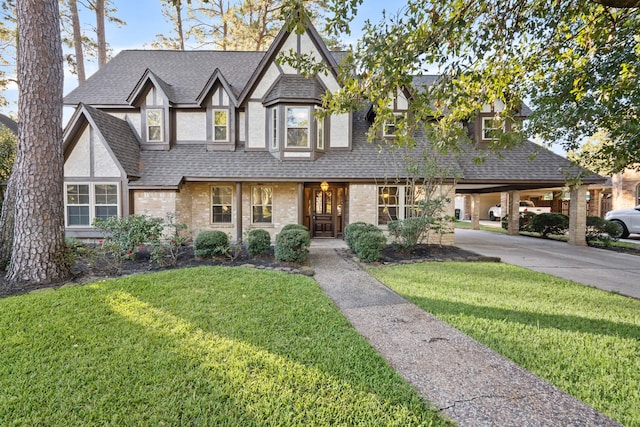 tudor-style house with brick siding, stucco siding, a shingled roof, a carport, and a front lawn