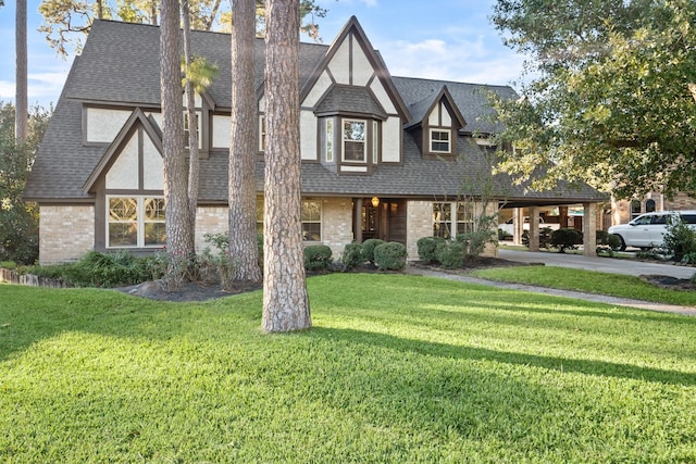 tudor-style house featuring stucco siding, roof with shingles, a front yard, a carport, and brick siding