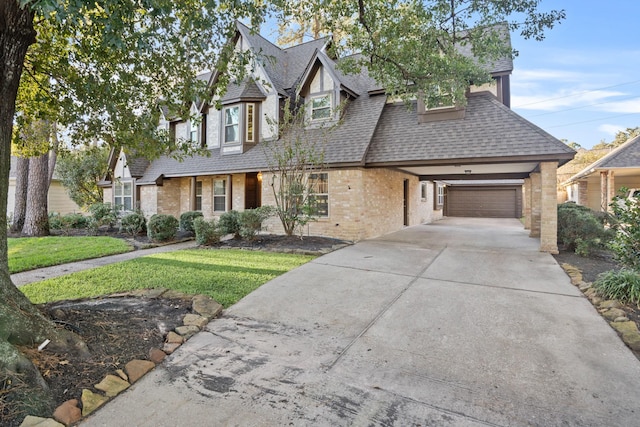 tudor home featuring driveway, a garage, roof with shingles, a front lawn, and brick siding