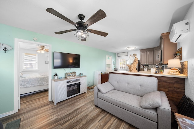 living room with a wall unit AC, a fireplace, wood finished floors, a ceiling fan, and baseboards