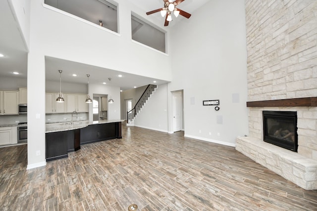 unfurnished living room with a stone fireplace, wood finished floors, a ceiling fan, baseboards, and stairway