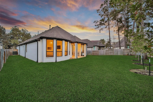 back of property at dusk with cooling unit, a fenced backyard, brick siding, a shingled roof, and a lawn