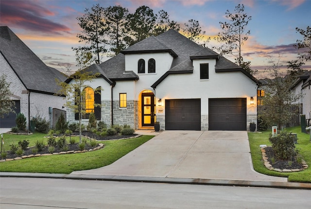 french country home featuring an attached garage, stone siding, a shingled roof, and a lawn