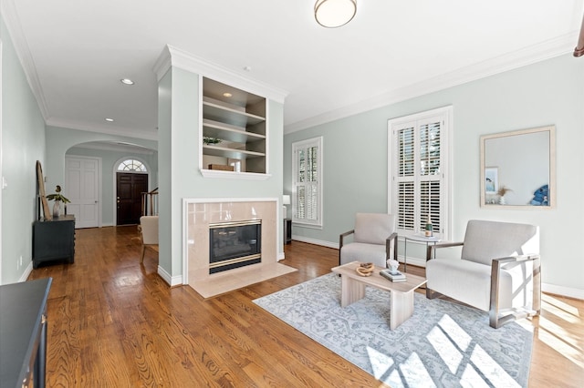 living room with baseboards, arched walkways, a tiled fireplace, ornamental molding, and wood finished floors