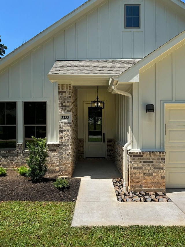 doorway to property with an attached garage, brick siding, board and batten siding, and roof with shingles