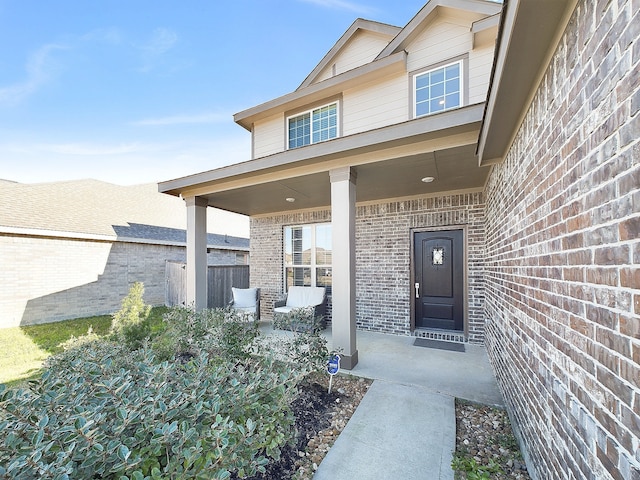 view of exterior entry featuring covered porch, brick siding, and fence