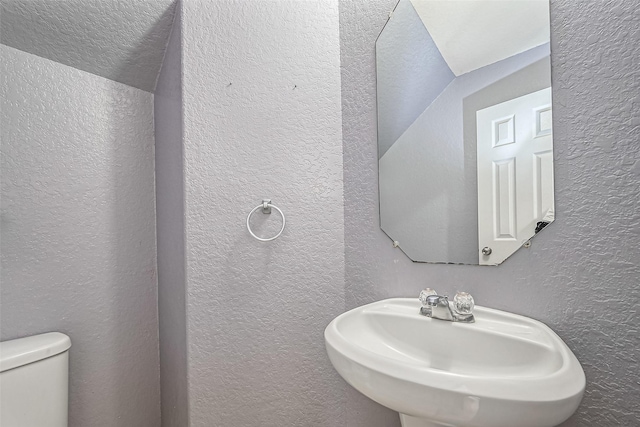 half bath with lofted ceiling, a textured wall, a sink, and toilet