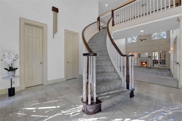 staircase featuring a towering ceiling, a warm lit fireplace, and baseboards