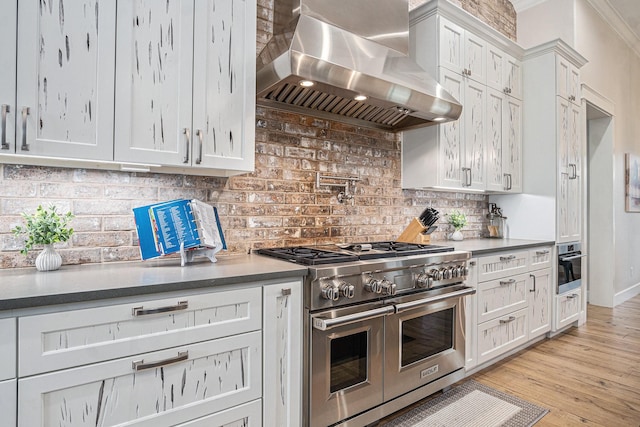 kitchen featuring stainless steel appliances, light wood-style floors, white cabinets, wall chimney range hood, and backsplash