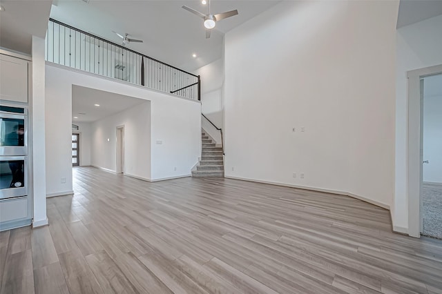 unfurnished living room featuring a high ceiling, light wood-style floors, ceiling fan, baseboards, and stairs