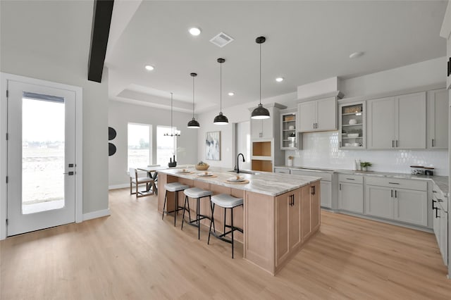 kitchen featuring a kitchen island with sink, a sink, light wood finished floors, a raised ceiling, and tasteful backsplash