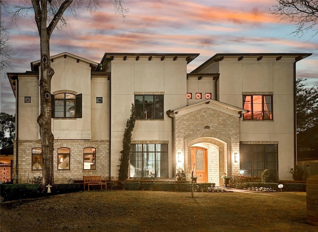 front of house at dusk featuring french doors, stone siding, and stucco siding