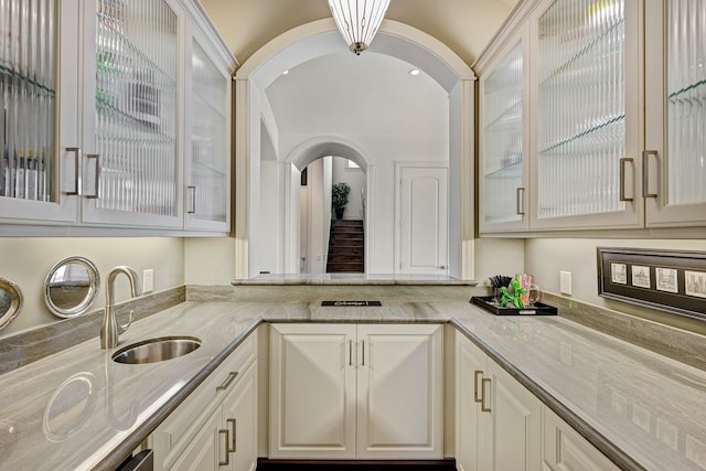 kitchen with light stone countertops, arched walkways, a sink, white cabinets, and glass insert cabinets