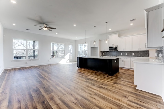 kitchen with stainless steel microwave, open floor plan, a kitchen island with sink, light countertops, and backsplash