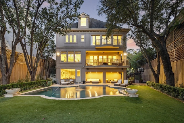 back of property at dusk with a balcony, a fenced backyard, outdoor lounge area, and a lawn