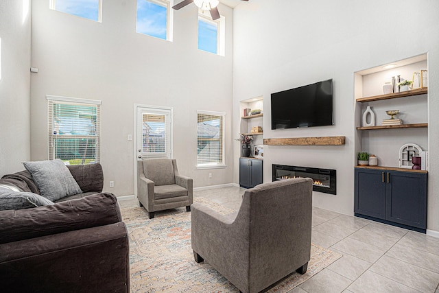 living area featuring built in shelves, a glass covered fireplace, light tile patterned flooring, ceiling fan, and baseboards