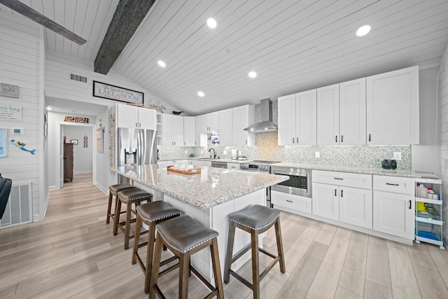 kitchen featuring a breakfast bar area, vaulted ceiling with beams, stainless steel appliances, light wood-type flooring, and wall chimney range hood