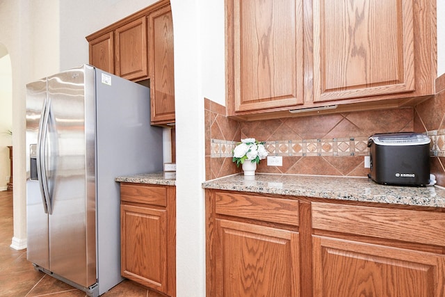 kitchen with light stone counters, arched walkways, stainless steel refrigerator with ice dispenser, backsplash, and brown cabinetry