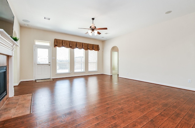 unfurnished living room featuring ceiling fan, arched walkways, a tile fireplace, dark wood-type flooring, and visible vents