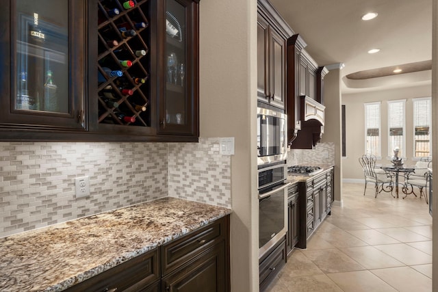 kitchen with dark brown cabinetry, light tile patterned floors, glass insert cabinets, appliances with stainless steel finishes, and light stone countertops