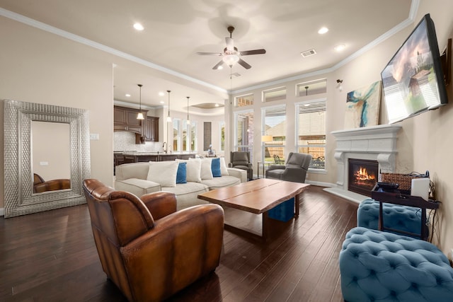 living area with dark wood-style floors, visible vents, ornamental molding, a ceiling fan, and a lit fireplace