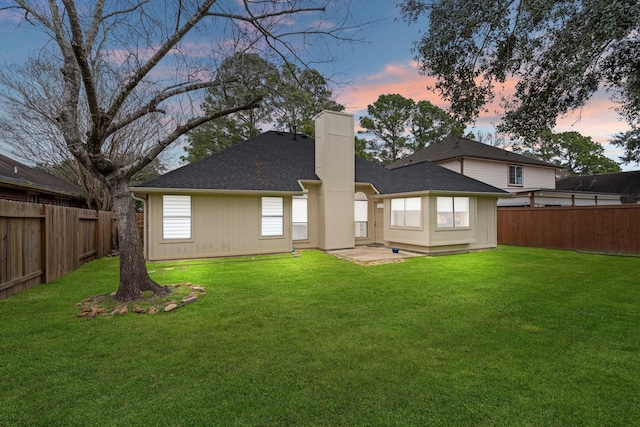 back of property at dusk with a fenced backyard, a chimney, roof with shingles, and a yard