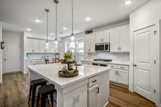 kitchen with a center island with sink, hanging light fixtures, appliances with stainless steel finishes, white cabinetry, and wood finished floors