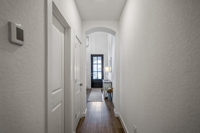 hall featuring baseboards, arched walkways, dark wood finished floors, and a textured wall