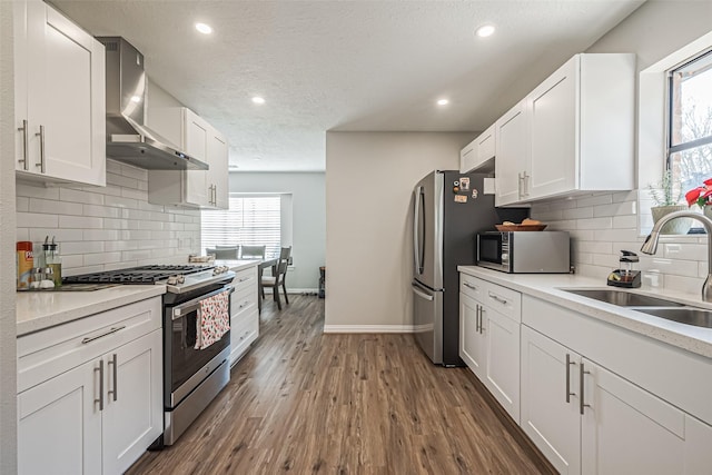 kitchen featuring dark wood-type flooring, wall chimney range hood, appliances with stainless steel finishes, white cabinetry, and a sink