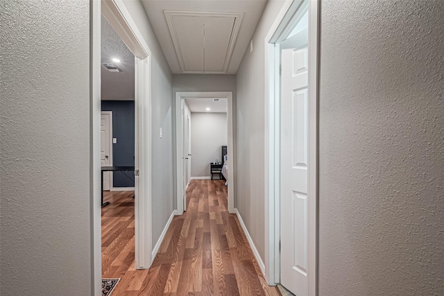 hall featuring baseboards, visible vents, light wood finished floors, attic access, and a textured wall
